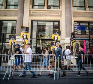 A group of striking workers holding signs stating “SAG-AFTRA ON STRIKE!” walking outside a brick building belonging to Warner Bros. Discovery. They are walking partially on the street and partially on the sidewalk, protected from cars by a mobile metal fence.