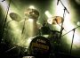 A drummer playing on stage with green overtones and light shining down on them. The snare drum is from Tama and reads “phones-music.com”.