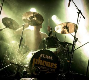 A drummer playing on stage with green overtones and light shining down on them. The snare drum is from Tama and reads “phones-music.com”.