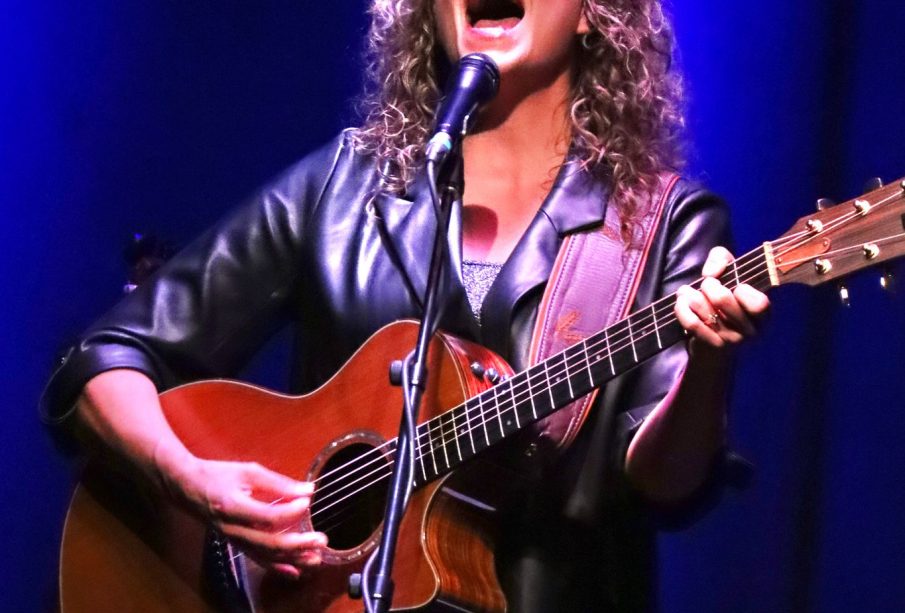 A woman with curly blonde hair is on a stage with a blue backdrop and a white light shining down on her. She is playing a wooden guitar and singing, her head thrown back with the force of her voice.