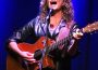 A woman with curly blonde hair is on a stage with a blue backdrop and a white light shining down on her. She is playing a wooden guitar and singing, her head thrown back with the force of her voice.