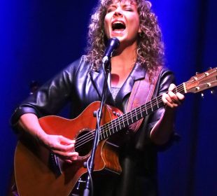 A woman with curly blonde hair is on a stage with a blue backdrop and a white light shining down on her. She is playing a wooden guitar and singing, her head thrown back with the force of her voice.