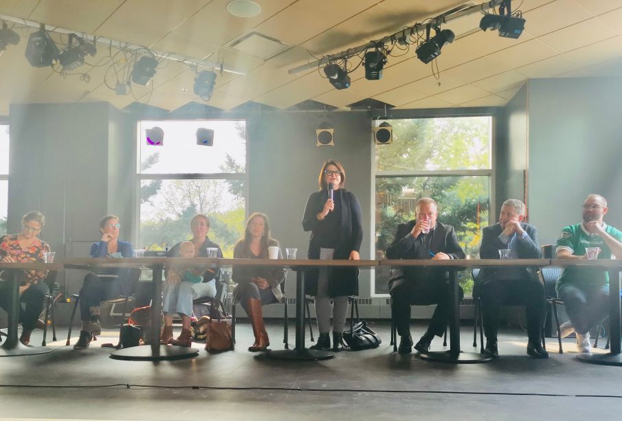 Eight political representatives range from left to right seated at tables facing the audience on stage at the UofR Lazy Owl. One representative near the middle is standing and speaking into a microphone