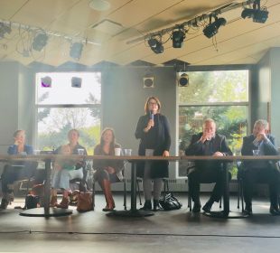 Eight political representatives range from left to right seated at tables facing the audience on stage at the UofR Lazy Owl. One representative near the middle is standing and speaking into a microphone