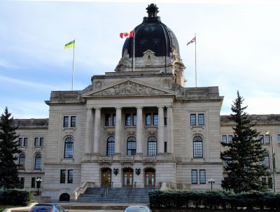The Saskatchewan Provincial Legislative Building fills the frame, a stone institutional building with a large dome at the centre of the roof and three flags flying from the roof against a white and light blue sky.