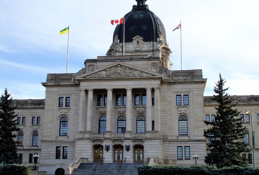 The Saskatchewan Provincial Legislative Building fills the frame, a stone institutional building with a large dome at the centre of the roof and three flags flying from the roof against a white and light blue sky.