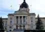 The Saskatchewan Provincial Legislative Building fills the frame, a stone institutional building with a large dome at the centre of the roof and three flags flying from the roof against a white and light blue sky.