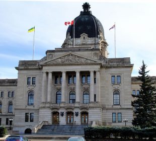 The Saskatchewan Provincial Legislative Building fills the frame, a stone institutional building with a large dome at the centre of the roof and three flags flying from the roof against a white and light blue sky.