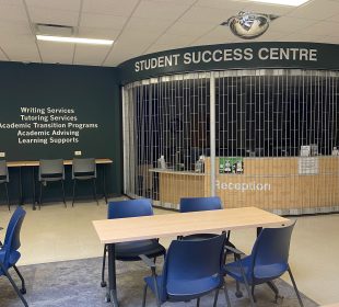 A banner of light-coloured text on a dark background titled Student Success Centre is above a reception area in an open room surrounded by study tables and blue chairs.