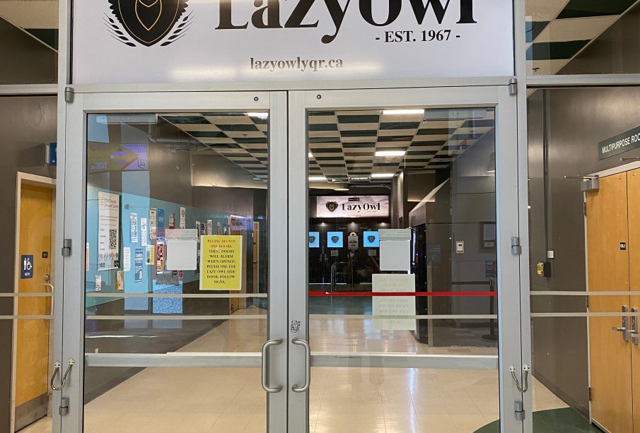 A photo of the double doors to the Lazy Owl at the University of Regina. The doors are made of glass and stainless steel. Above the doors there is a white banner containing a line art image of an owl in a shield and “LazyOwl” in bold, black lettering. Beneath the lettering is “EST. 1967” and the Lazy Owl’s website link: lazyowlyqr.ca.