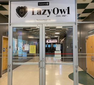 A photo of the double doors to the Lazy Owl at the University of Regina. The doors are made of glass and stainless steel. Above the doors there is a white banner containing a line art image of an owl in a shield and “LazyOwl” in bold, black lettering. Beneath the lettering is “EST. 1967” and the Lazy Owl’s website link: lazyowlyqr.ca.