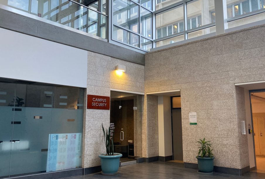 This photo shows a red sign with grey lettering reading “Campus Security” mounted on a wall to the left of an interior glass door in an austere institutional building of glass and Tyndall Stone.
