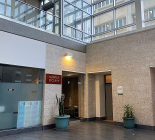 This photo shows a red sign with grey lettering reading “Campus Security” mounted on a wall to the left of an interior glass door in an austere institutional building of glass and Tyndall Stone.