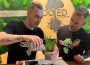 Two people with short blond hair shaved on the sides of their heads and shirts denoting the store they work at, Rooted Living Designs, are adding soil to a plant pot with two cacti in it. The wall behind the people is covered in moss and has railroad ties behind a circle of wood that has been carved to reflect the store’s name with the image of a tree with long roots as the “t” in the word “Rooted”.