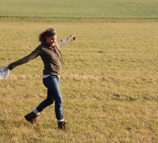 A person standing in an open field, arms stretched. They are holding a Frisbee in one hand.