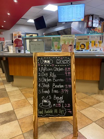 A photo of the entrance to Gong-Cha and its food menu. The menu is a blackboard written on with various colours of chalk, titled “Food Menu” with a smile drawn under the two “o”s in “food”. It contains items such as popcorn chicken for $8.99, dry ribs for $9.99, a chicken burrito $3.99, steak bourbon for $5.99, french fries for $3.99, and a medium brewed coffee for $1.95 or a large for $2.25. There is a drawing of a mug of coffee releasing steam in the shape of a heart and a cloud with “Food available all day” written below it and beside that it says “+ tax,”underlined twice.
