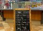A photo of the entrance to Gong-Cha and its food menu. The menu is a blackboard written on with various colours of chalk, titled “Food Menu” with a smile drawn under the two “o”s in “food”. It contains items such as popcorn chicken for $8.99, dry ribs for $9.99, a chicken burrito $3.99, steak bourbon for $5.99, french fries for $3.99, and a medium brewed coffee for $1.95 or a large for $2.25. There is a drawing of a mug of coffee releasing steam in the shape of a heart and a cloud with “Food available all day” written below it and beside that it says “+ tax,”underlined twice.