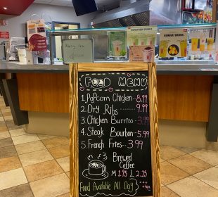 A photo of the entrance to Gong-Cha and its food menu. The menu is a blackboard written on with various colours of chalk, titled “Food Menu” with a smile drawn under the two “o”s in “food”. It contains items such as popcorn chicken for $8.99, dry ribs for $9.99, a chicken burrito $3.99, steak bourbon for $5.99, french fries for $3.99, and a medium brewed coffee for $1.95 or a large for $2.25. There is a drawing of a mug of coffee releasing steam in the shape of a heart and a cloud with “Food available all day” written below it and beside that it says “+ tax,”underlined twice.