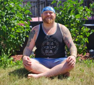 Yoga instructor Sacha Wolfson sits cross-legged outside with his hands on his knees, enjoying the sunshine.