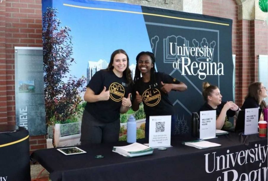 Two people standing at a registration table smiling at the camera. Two others are sitting, talking to someone off camera.