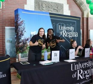 Two people standing at a registration table smiling at the camera. Two others are sitting, talking to someone off camera.