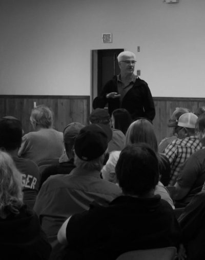 in a black and white photograph a white-haired male addresses a group of seated individuals seen from behind