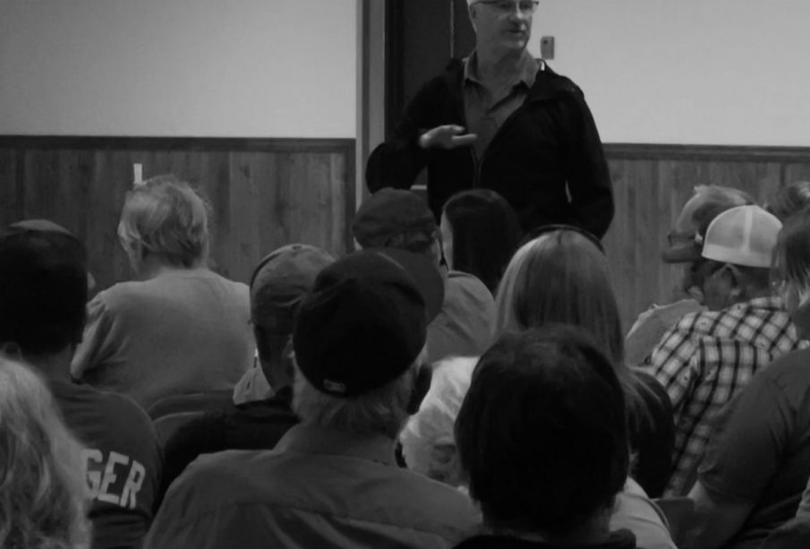 in a black and white photograph a white-haired male addresses a group of seated individuals seen from behind