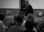 in a black and white photograph a white-haired male addresses a group of seated individuals seen from behind