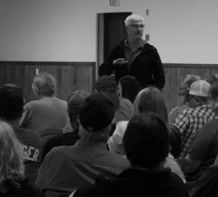 in a black and white photograph a white-haired male addresses a group of seated individuals seen from behind