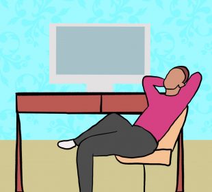 A person in black pants and a hot pink sweater sits in an office chair, relaxing in front of their desk with their computer turned off.