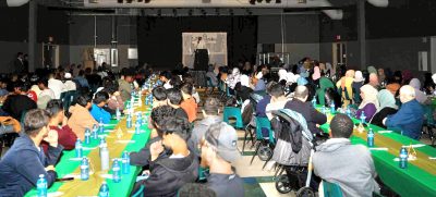 A photo shows people seated at banquet tables that fill a large room with a speaker facing the crowd from a stage at the furthest point in the photo.