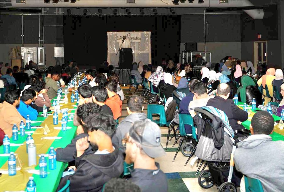 A photo shows people seated at banquet tables that fill a large room with a speaker facing the crowd from a stage at the furthest point in the photo.