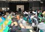 A photo shows people seated at banquet tables that fill a large room with a speaker facing the crowd from a stage at the furthest point in the photo.