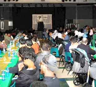 A photo shows people seated at banquet tables that fill a large room with a speaker facing the crowd from a stage at the furthest point in the photo.