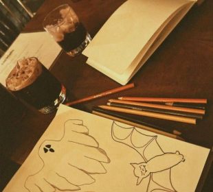 A photo of a table with two art filled notebooks (one near the camera and the other further down the table), coloured pencils strewn across the table, two dark beverages with ice in whiskey glasses in the middle of the table, and a deep red plastic cup at the far end of the table. The closer notebook has drawings of a ghost, a winking jack-o’-lantern, and a smiling bat while the further notebook has an unfinished drawing of a bat.