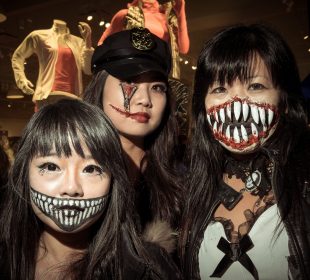 Three people with Halloween themed make-up in a photograph. The one on the left side has drawn a smiling mouth with sharp teeth onto her face, the one in the middle has a zipper opening her cheek under her eye and a scar pulling the side of her mouth open by using liquid latex, and the one on the right has a liquid latex and fake sharp tooth-ed mouth stretching across her face and covering her nose.