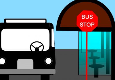 Looking at the front of a city bus stopped at a glass-enclosed bus stop with a red sign with white text spelling “Bus Stop.”