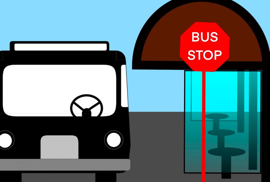 Looking at the front of a city bus stopped at a glass-enclosed bus stop with a red sign with white text spelling “Bus Stop.”