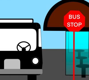Looking at the front of a city bus stopped at a glass-enclosed bus stop with a red sign with white text spelling “Bus Stop.”