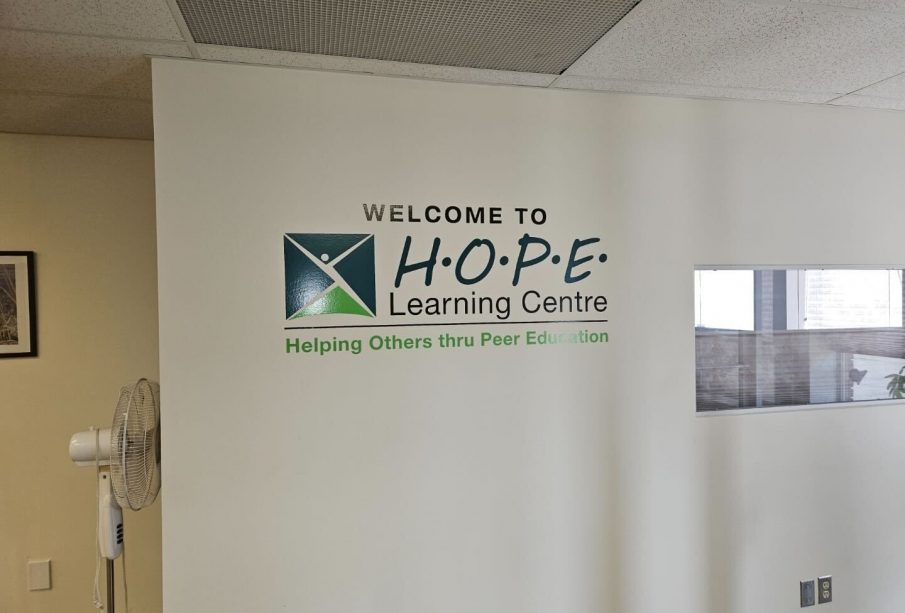 A white wall with the phrase “Welcome to HOPE Learning Centre” in black words with the phrase “Helping Others thru Peer Education” beneath it.