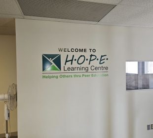 A white wall with the phrase “Welcome to HOPE Learning Centre” in black words with the phrase “Helping Others thru Peer Education” beneath it.