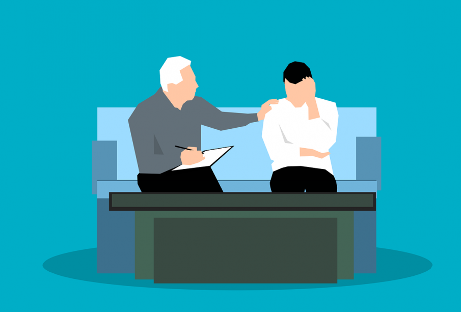 A young adult is sitting on a couch with a counsellor, who has their hand on their shoulder supportively.