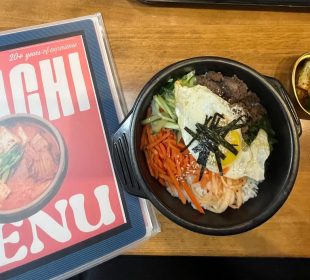 A photo of the front of a menu that reads “Kimchi Menu” and a meal. The meal consists of banchan in a stone bowl and a side of seasoned cucumbers and apple slices.