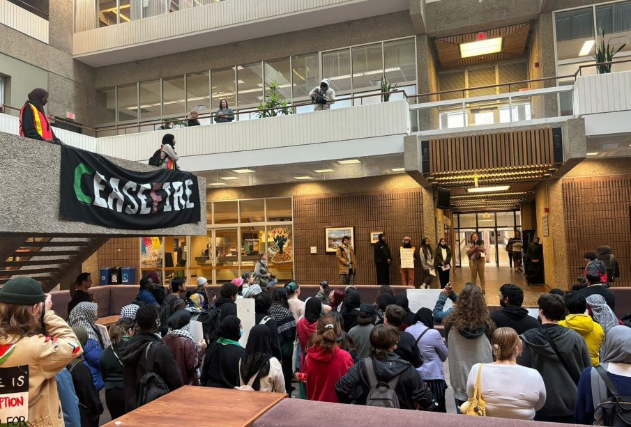 Protestors gather inside to listen to speakers with a “Cease Fire” sign hanging from an upper level.