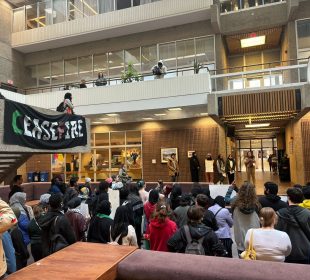 Protestors gather inside to listen to speakers with a “Cease Fire” sign hanging from an upper level.