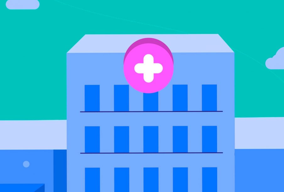 A blue hospital with a pink and white emergency cross sign above the entrance with a teal sky in the background.