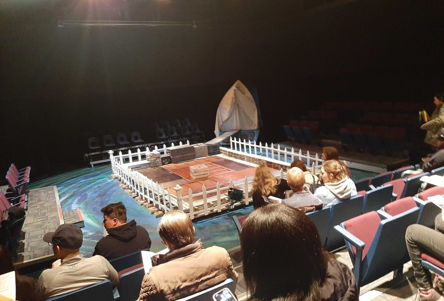 A photo of a stage set up with a fence around it, a gangway to the back right corner leading into a sheet doorway, and various trunks around the gangway and inside the fence.