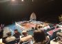 A photo of a stage set up with a fence around it, a gangway to the back right corner leading into a sheet doorway, and various trunks around the gangway and inside the fence.