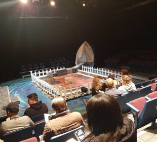 A photo of a stage set up with a fence around it, a gangway to the back right corner leading into a sheet doorway, and various trunks around the gangway and inside the fence.