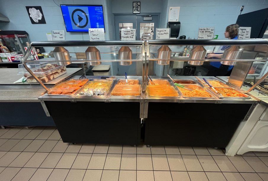 A buffet of 11 food choices with a bucket of buns off to the side. There is a server behind the buffet.
